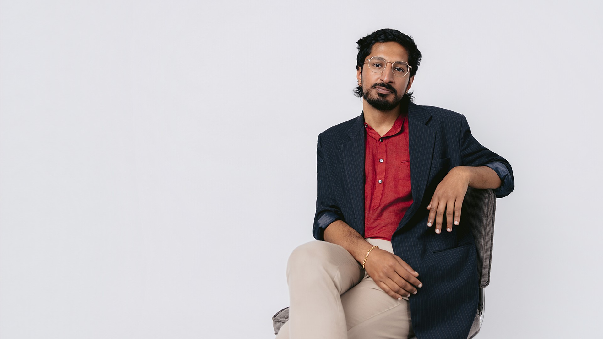 Arun sitzt mit übereinandergeschlagenen Beinen auf einem Stuhl, trägt ein rotes Hemd, ein dunkles Sakko, eine Brille und helle Hose und blickt vor hellem Hintergrund in die Kamera.