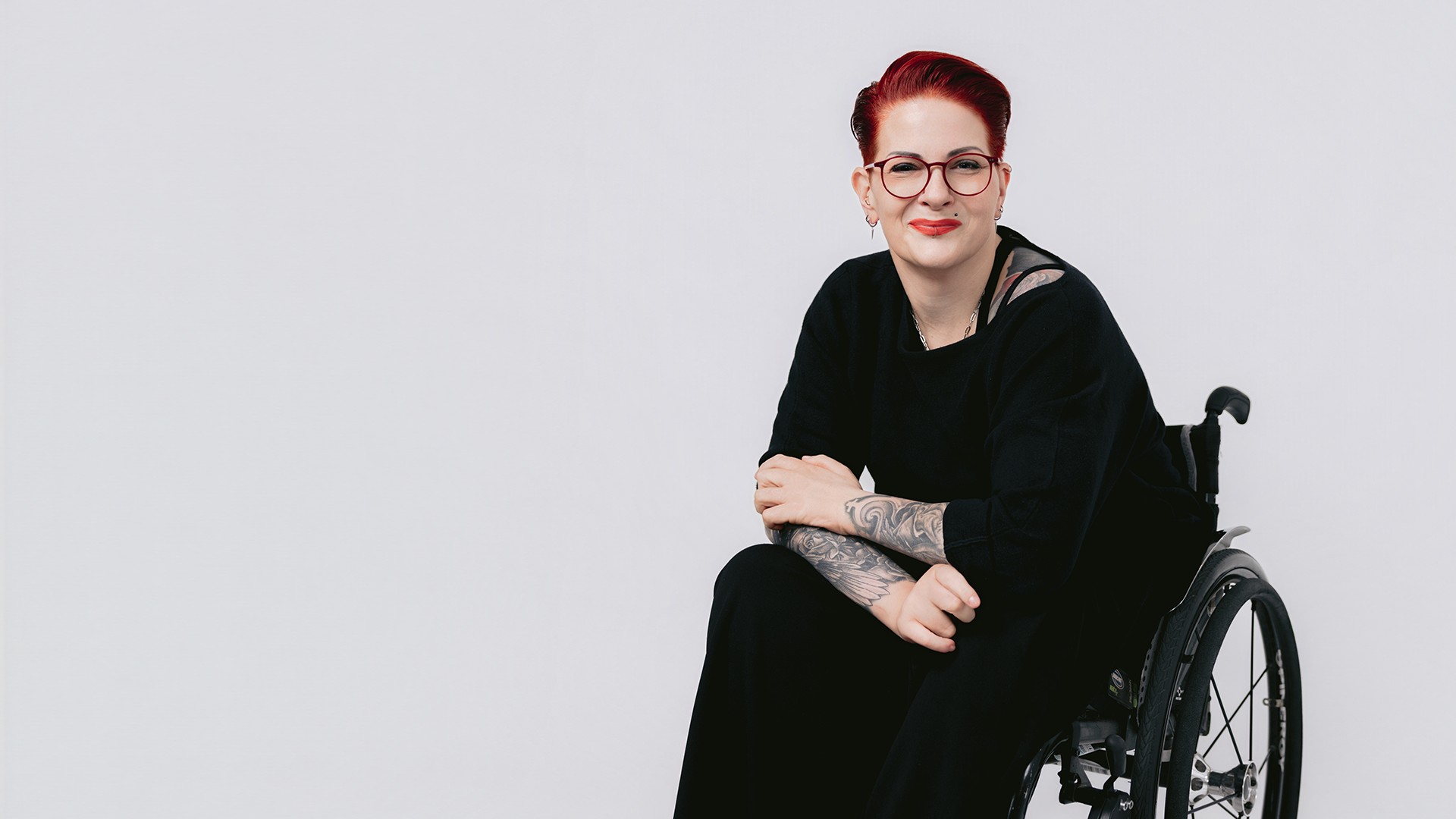 Claudia sitzt in einem Rollstuhl, trägt schwarze Kleidung und eine runde Brille, hat kurzes rotes Haar und tätowierte Unterarme und blickt vor hellem Hintergrund in die Kamera.