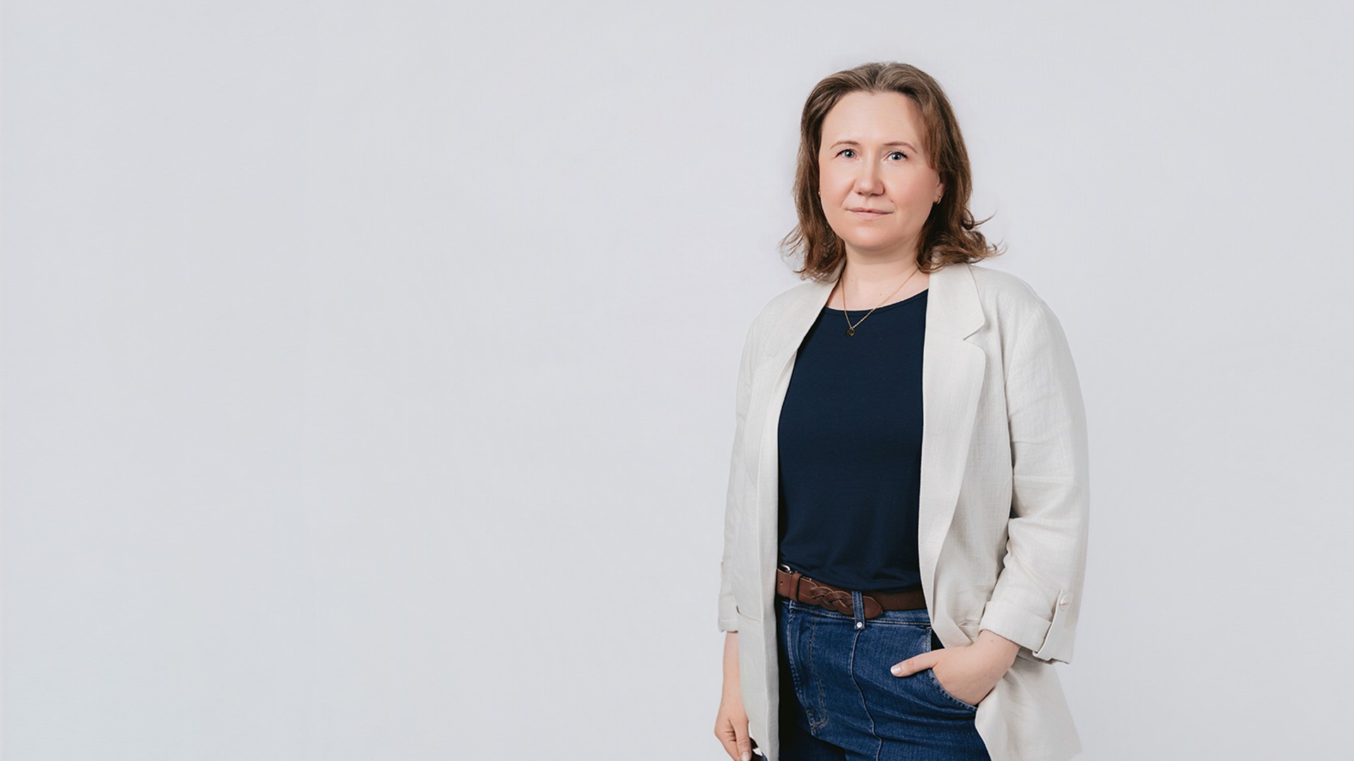 Desiree steht vor hellem Hintergrund, trägt ein dunkles Oberteil, einen hellen Blazer und eine Jeans, hat eine Hand in der Hosentasche und blickt in die Kamera.