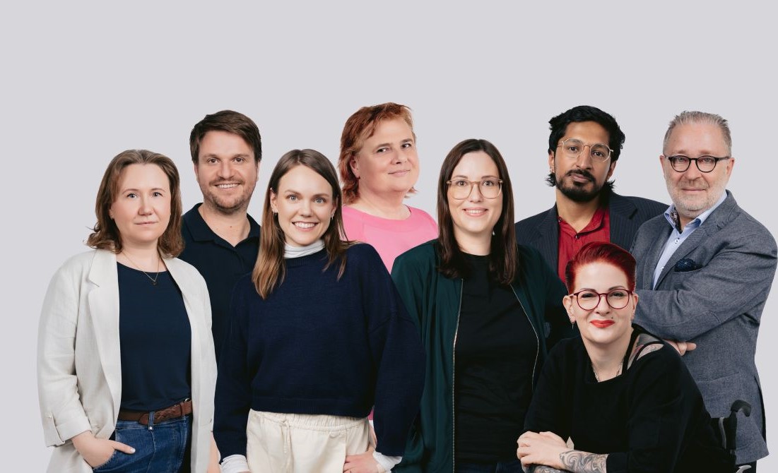 Acht Personen stehen und sitzen gruppiert vor hellem Hintergrund und blicken in die Kamera.