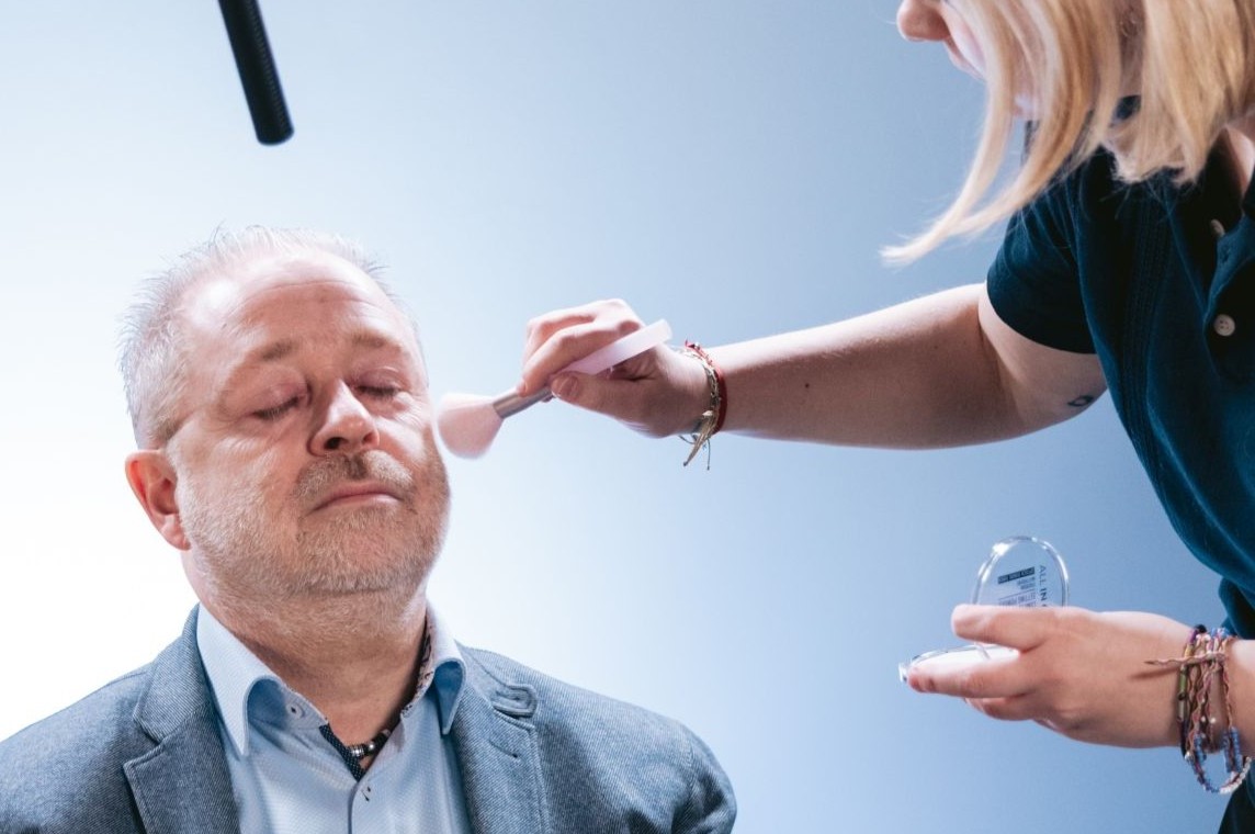 Person in grauem Sakko sitzt in einem Filmstudio, während eine weitere Person mit einem Pinsel Make-up aufträgt; über ihnen ist ein Mikrofon an einer Tonangel positioniert.