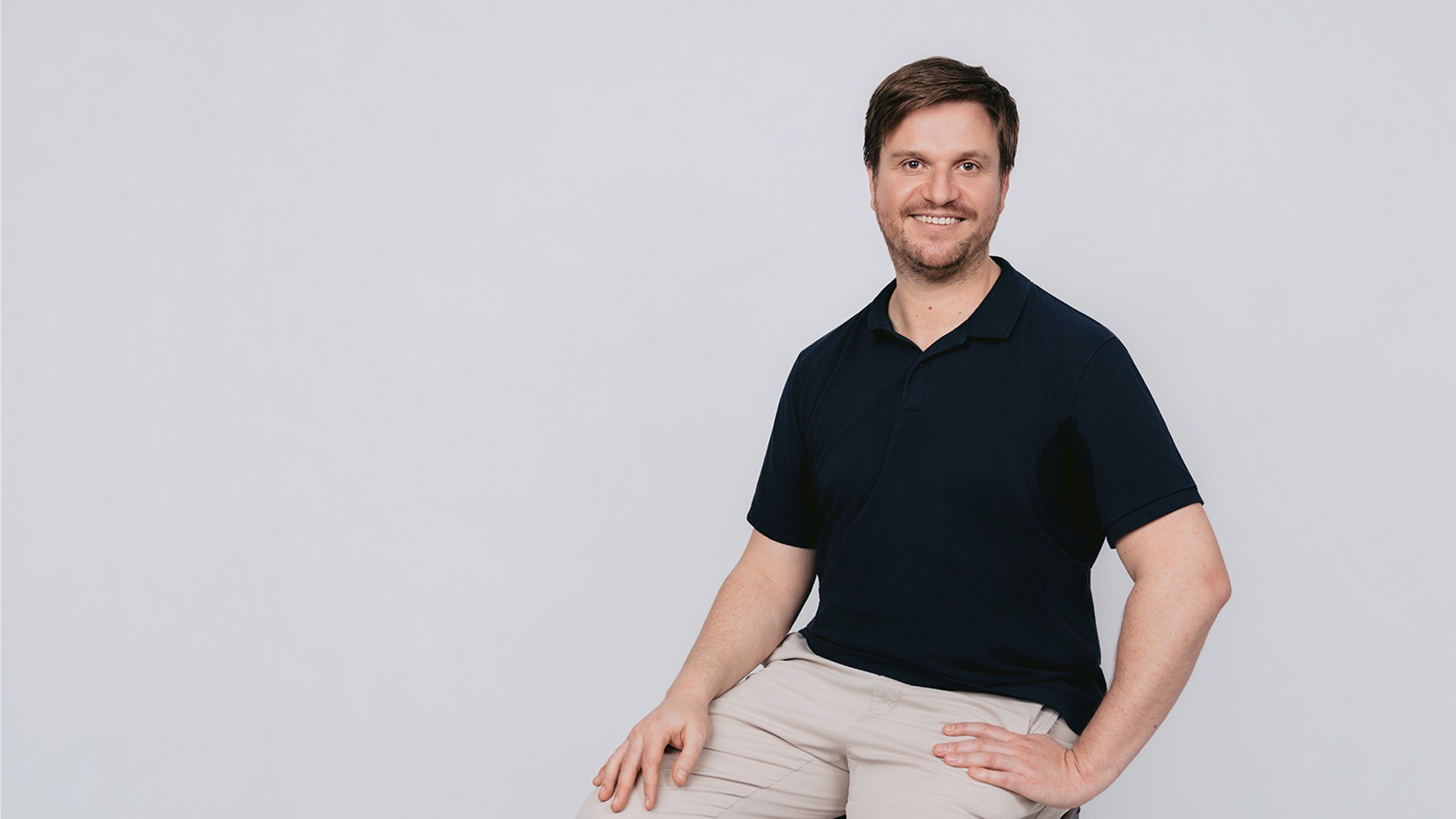 Michael sitzt auf einem Hocker, trägt ein dunkles Poloshirt und helle Hose und blickt lächelnd vor hellem Hintergrund in die Kamera.