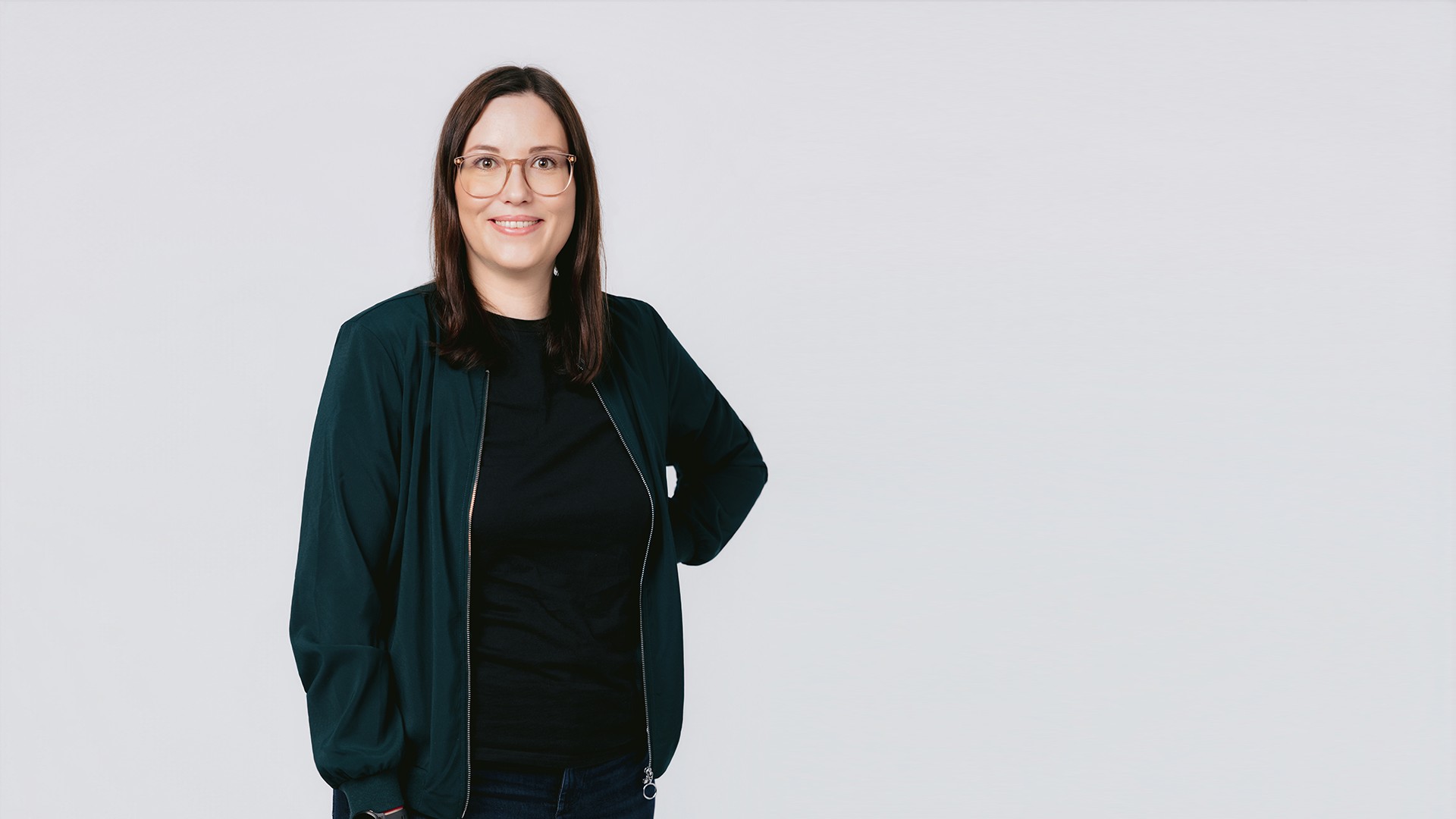 Susi steht vor hellem Hintergrund, trägt eine Brille, eine dunkle Jacke über einem schwarzen Oberteil sowie eine Jeans und blickt lächelnd in die Kamera.