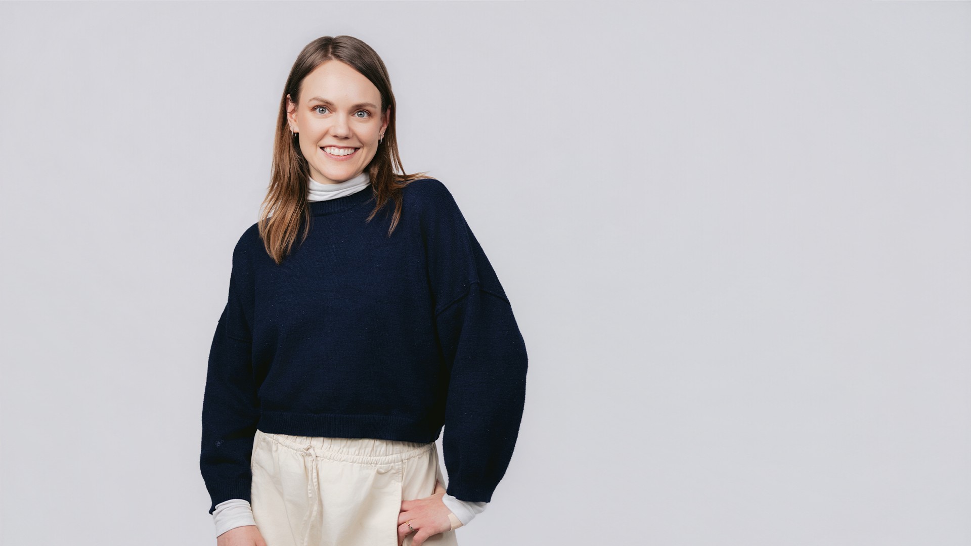 Vera steht vor hellem Hintergrund, trägt einen dunklen Pullover über einem hellen Oberteil und eine helle Hose, hat eine Hand an der Hüfte und blickt lächelnd in die Kamera.