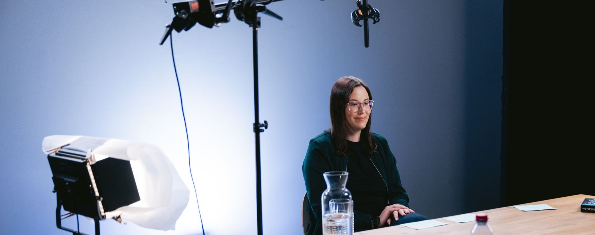 Person mit Brille sitzt an einem Tisch in einem Filmstudio, umgeben von Kameras, Mikrofonen, Scheinwerfern und weiterem Aufnahmeequipment.
